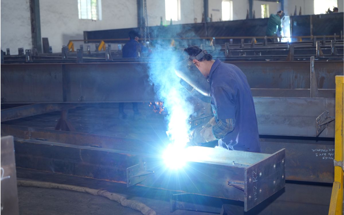 A worker wearing a welding helmet and blue uniform welds a heavy steel I-beam inside a fabrication facility, creating a bright blue arc, smoke, and sparks.